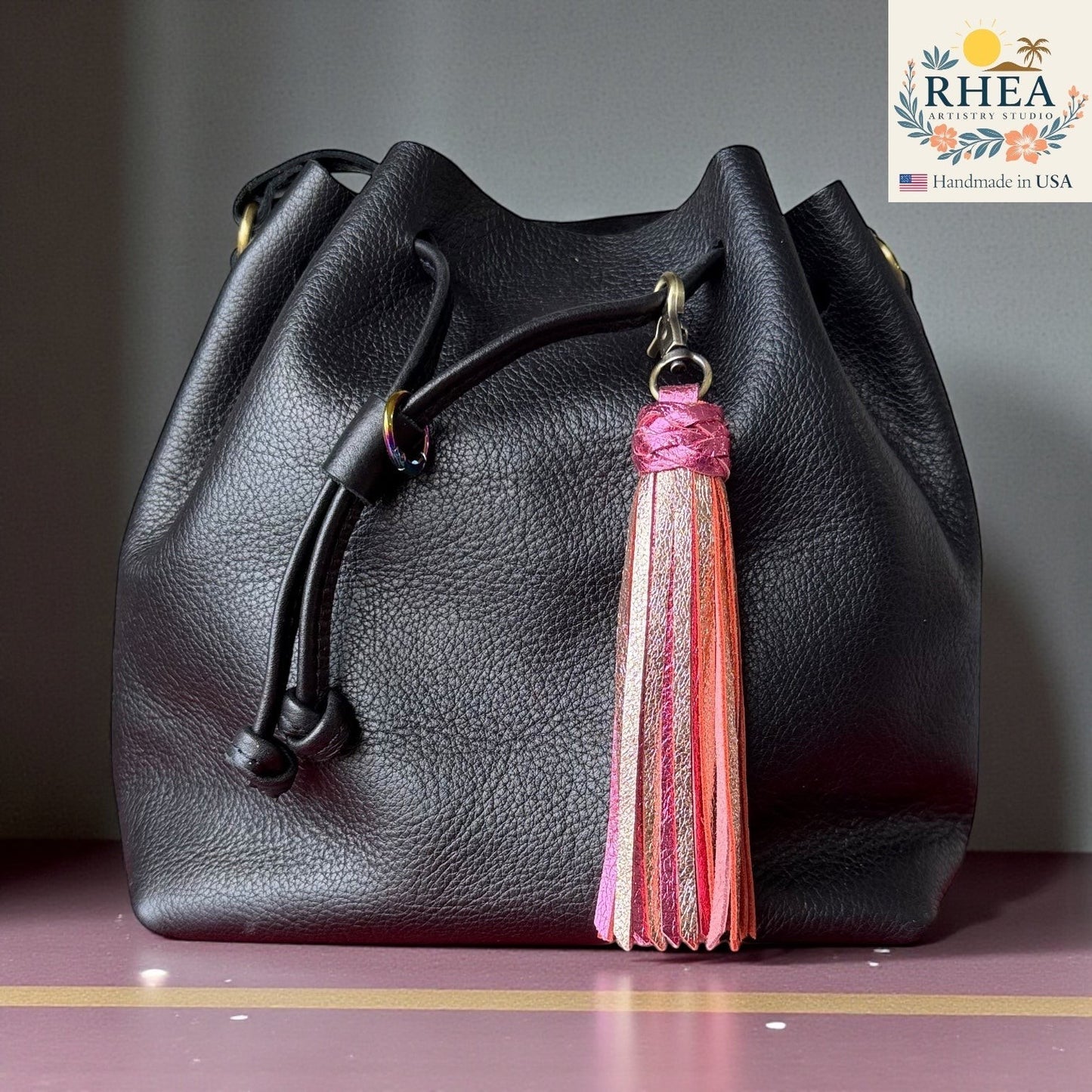 Rhea Artistry Studio handmade leather tassel charm with matte and metallic pink, coral, and gold finishes attached to a black leather bag.