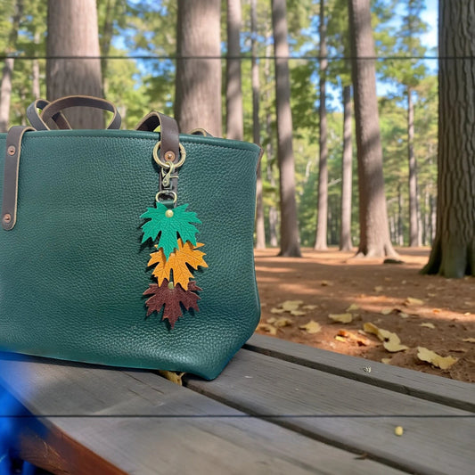 Rhea Artistry Studio layered maple leaf leather bag charm in green, orange, and brown with vein detail, attached to a dark green tote bag.