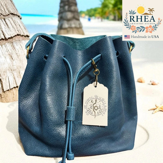 Rhea Artistry Studio navy blue Saffiano leather drawstring bucket bag with engraved Empower Collection tag on a beach.
