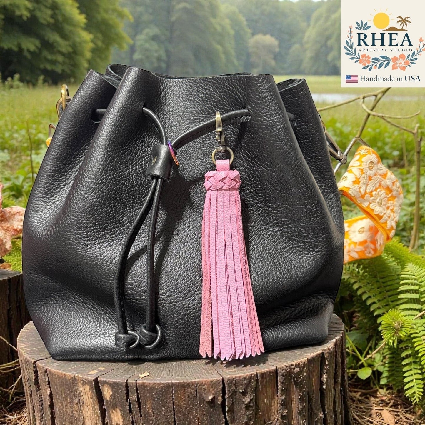 Rhea Artistry Studio pink leather tassel bag charm with braided detail and brass clasp, attached to a black leather bucket bag.