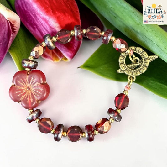 Rhea Artistry Studio handmade bracelet with red glass beads, floral focal bead, and brass toggle clasp, set against tulips.