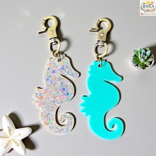 Rhea Artistry Studio holographic glitter and teal acrylic seahorse charms with brass clasps. Coastal-themed bag charms or zipper pulls.