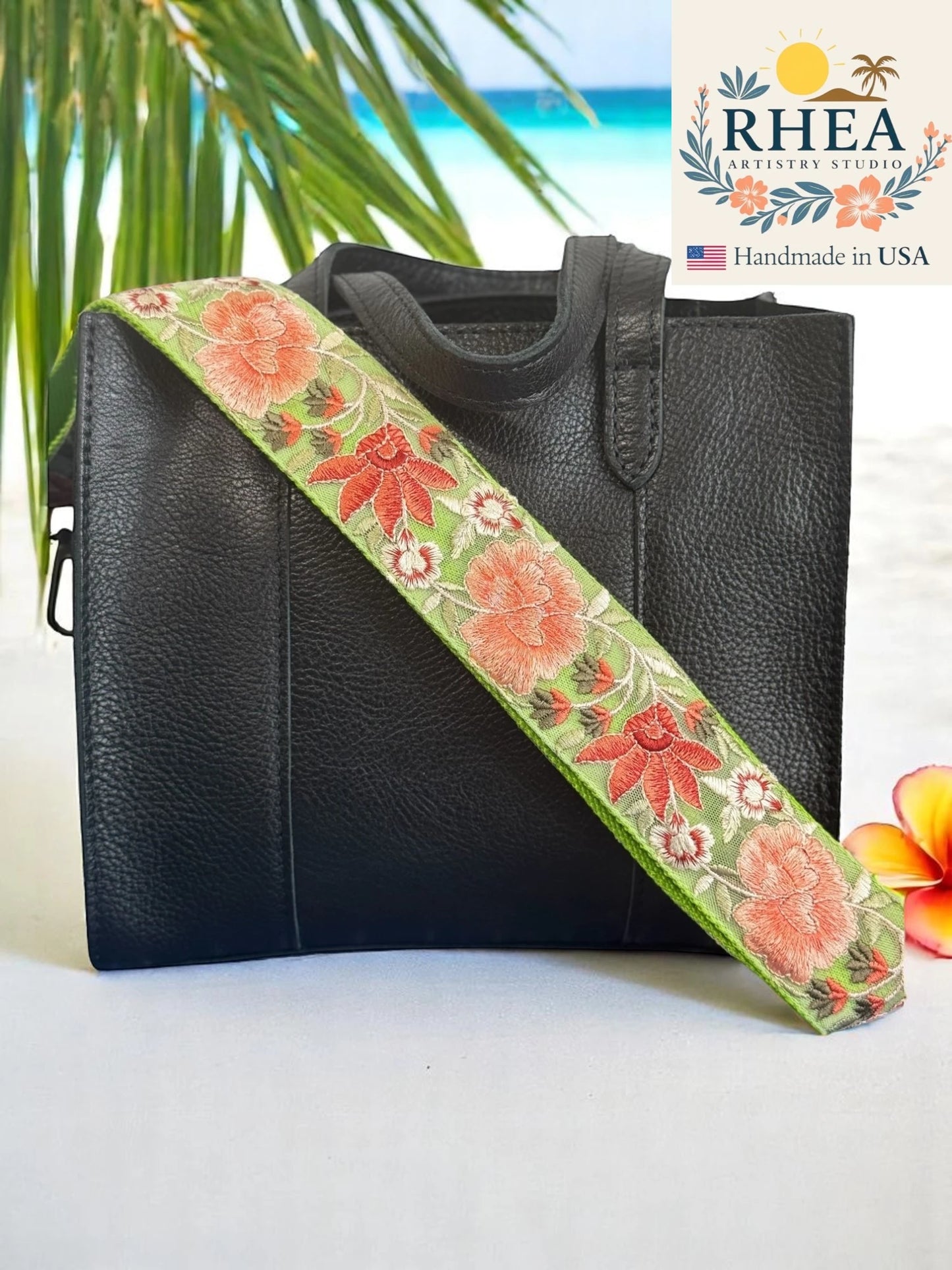 Black leather handbag with a floral purse strap on a white surface, palm leaves, and RHEA branding in the background.