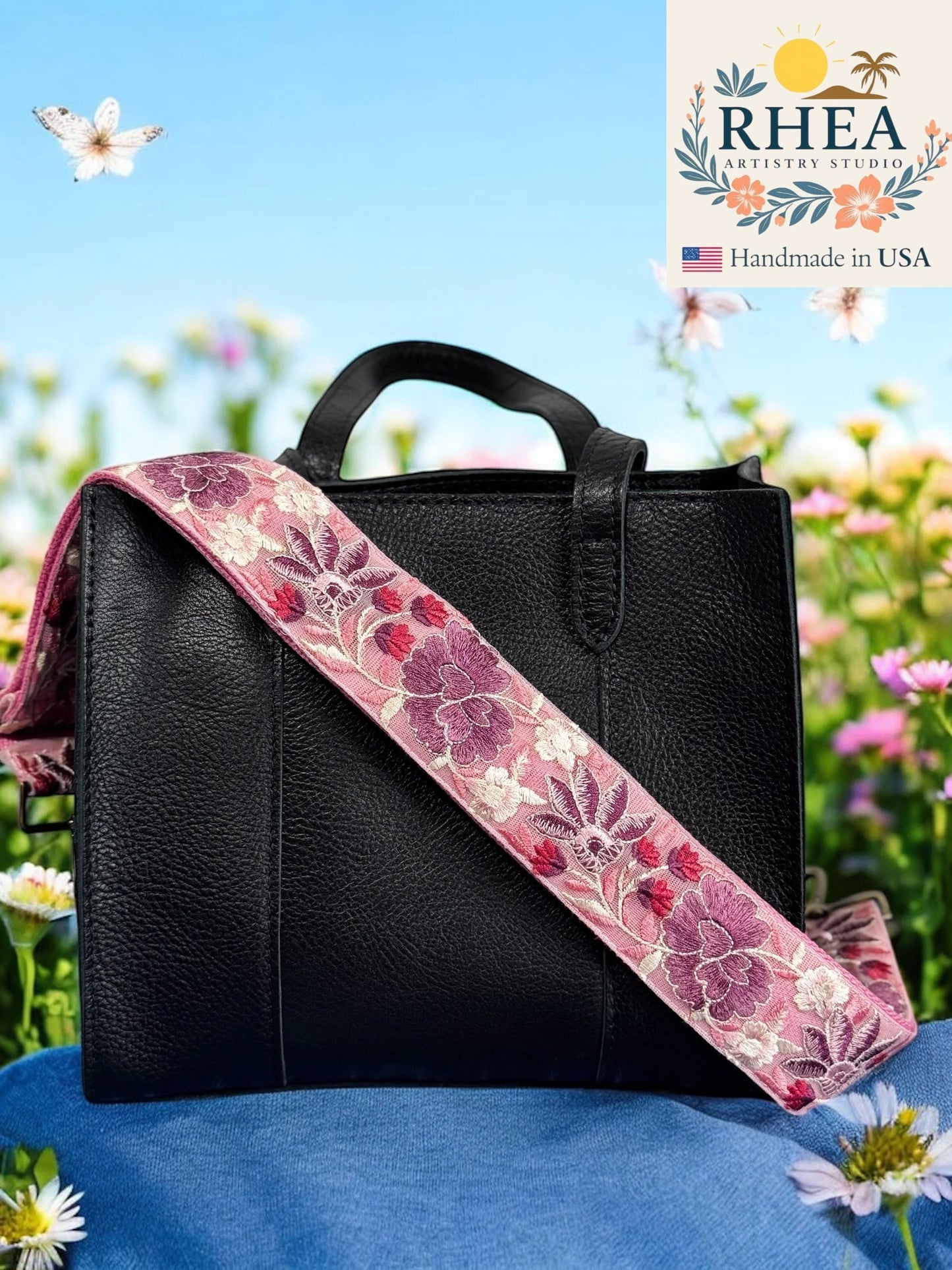 Black handbag with a floral purse strap on a blue surface with flowers in the background, featuring the RHEA Artisans Studio logo.