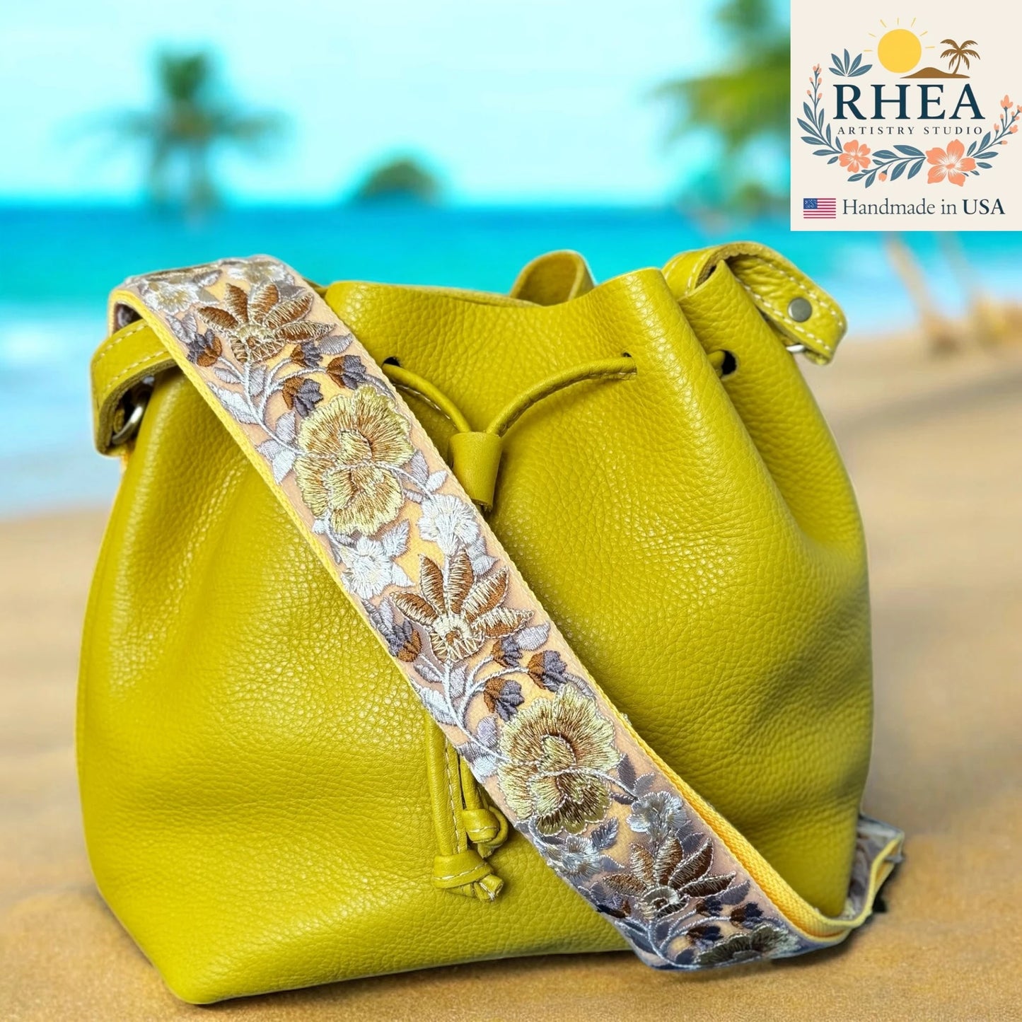 Yellow leather bag with floral purse strap on a beach background, featuring RHEA Artistry Studio logo.