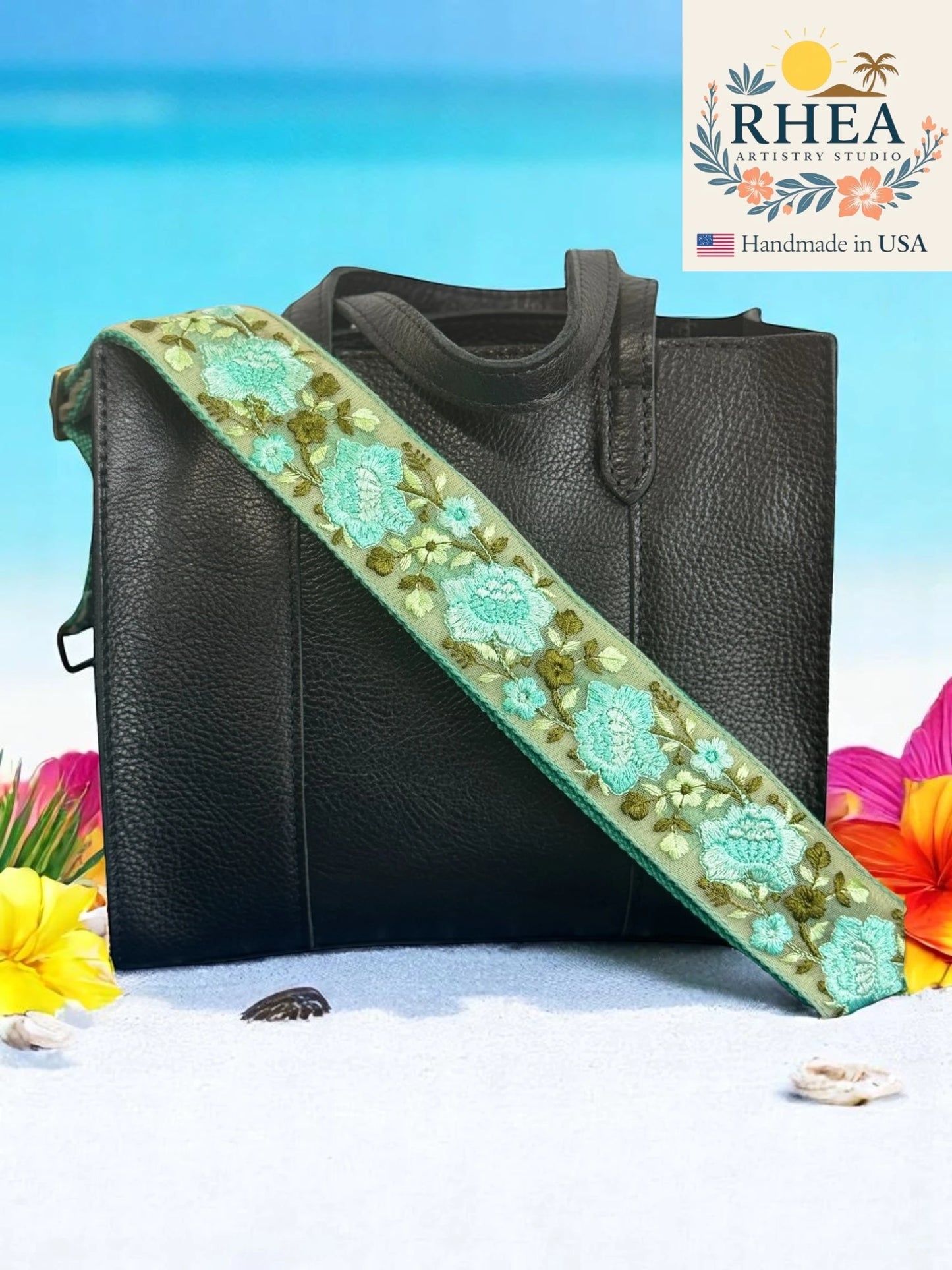 Black handbag with a floral purse strap against a beach background, featuring the RHEA brand.
