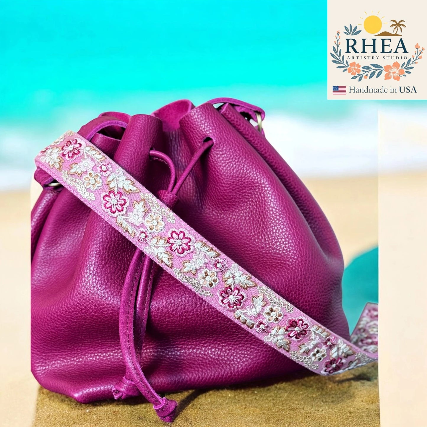 Pink leather drawstring bag with floral strap on a beach background, featuring the RHEA brand.
