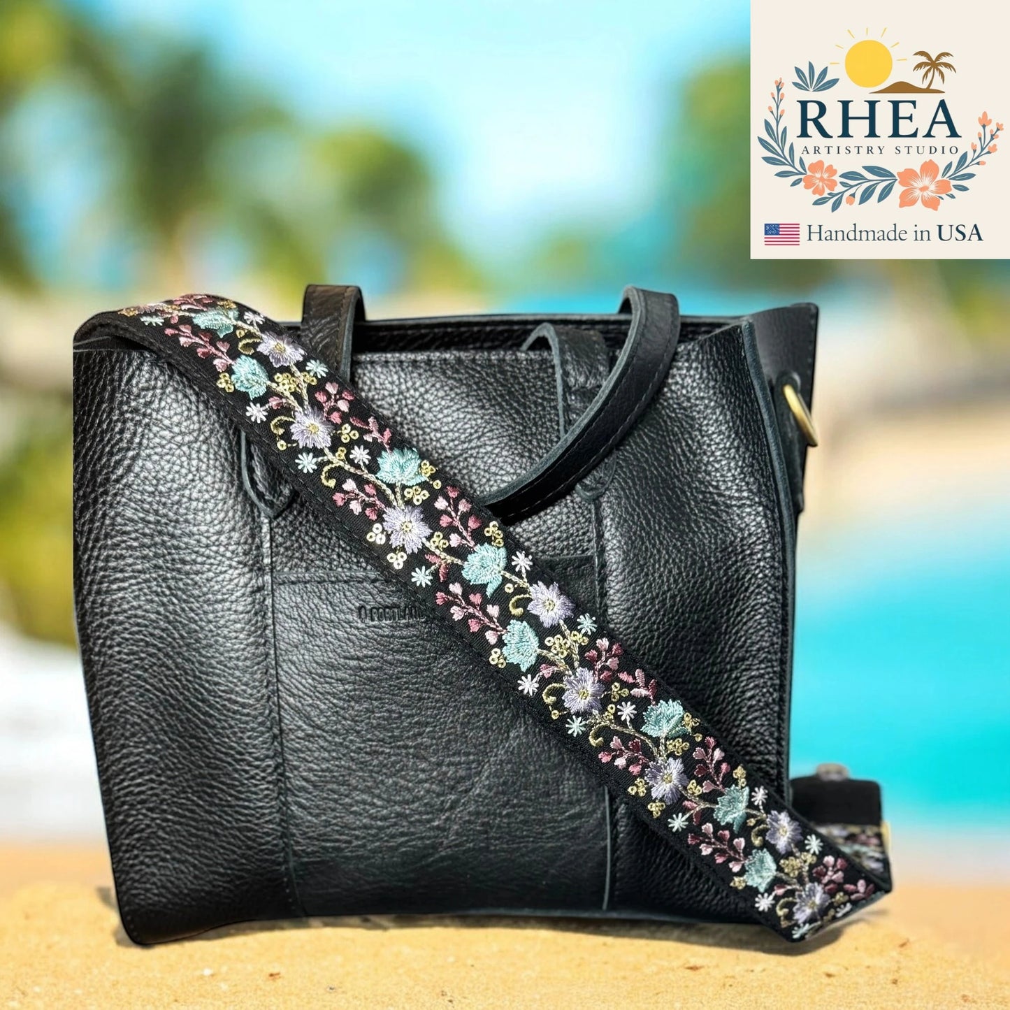 Black handbag with floral strap on a beach background, featuring the RHEA Artistry Studio logo.