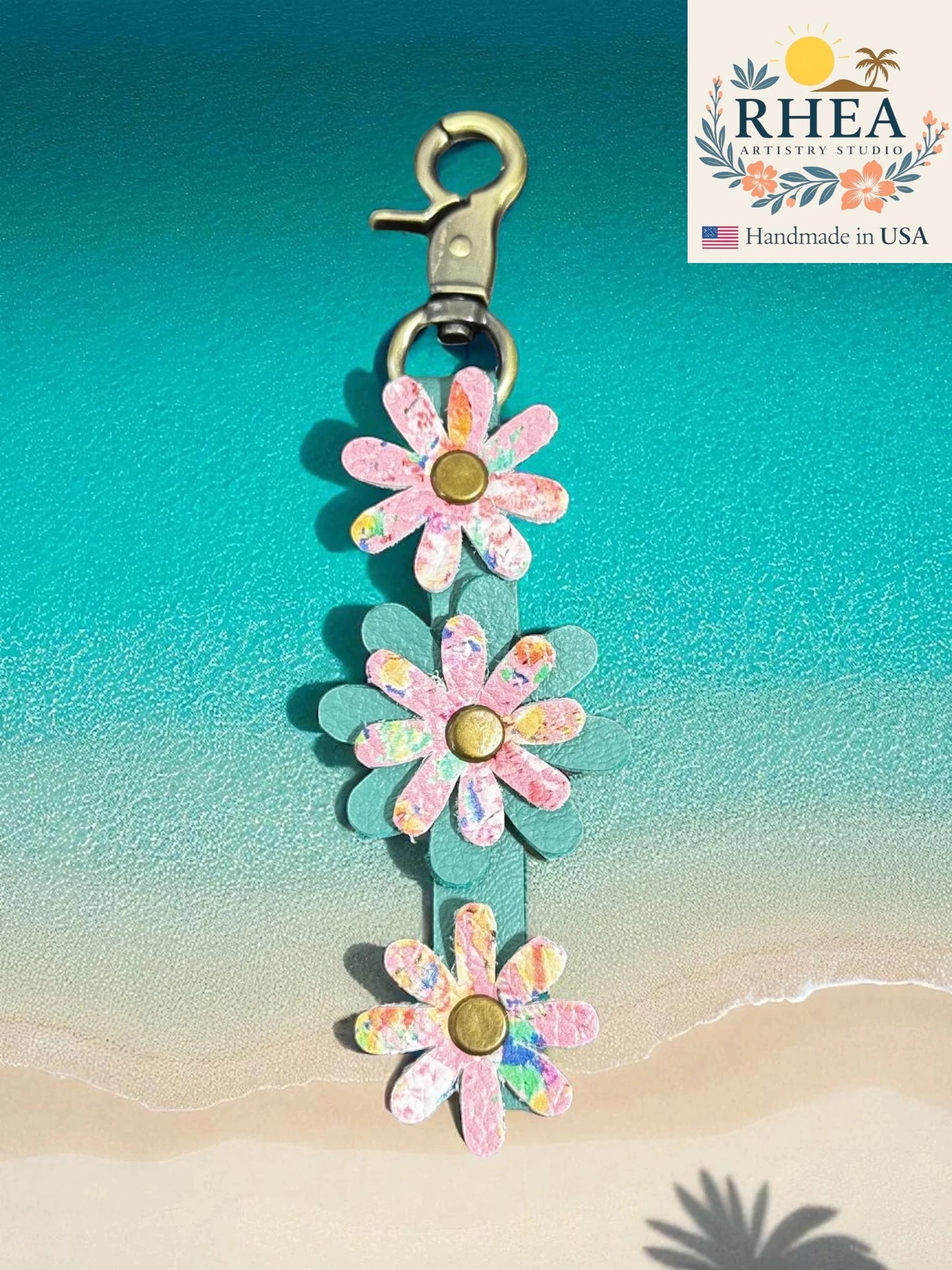 Flower-shaped leather purse charm with a clip on a beach background, branded 'RHEA'.