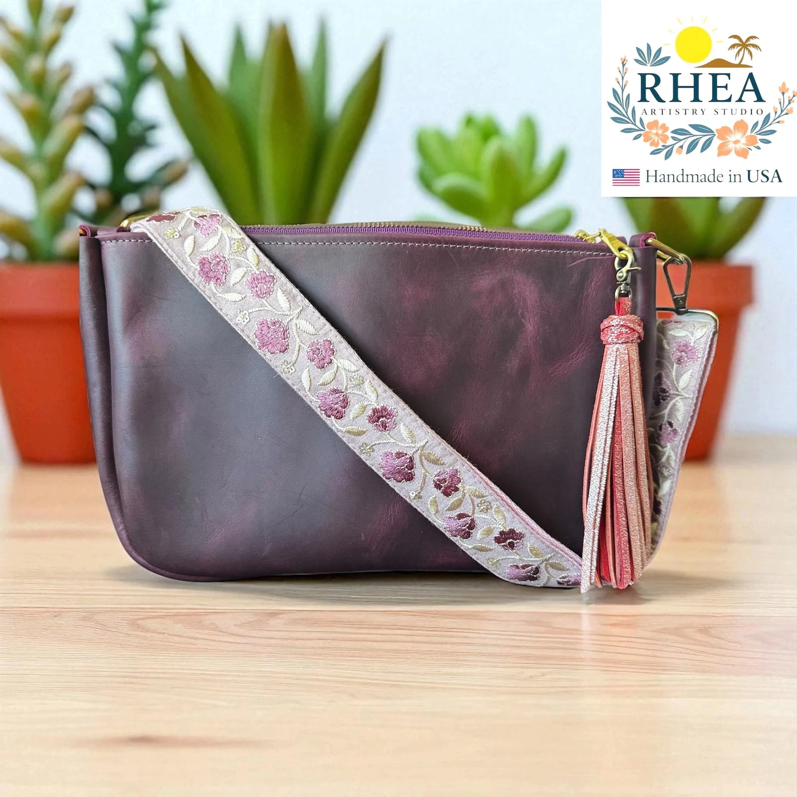 Purple handbag with floral leather purse strap and tassel on a wooden surface, RHEA brand visible.