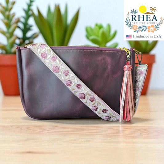 Purple handbag with floral leather purse strap and tassel on a wooden surface, RHEA brand visible.