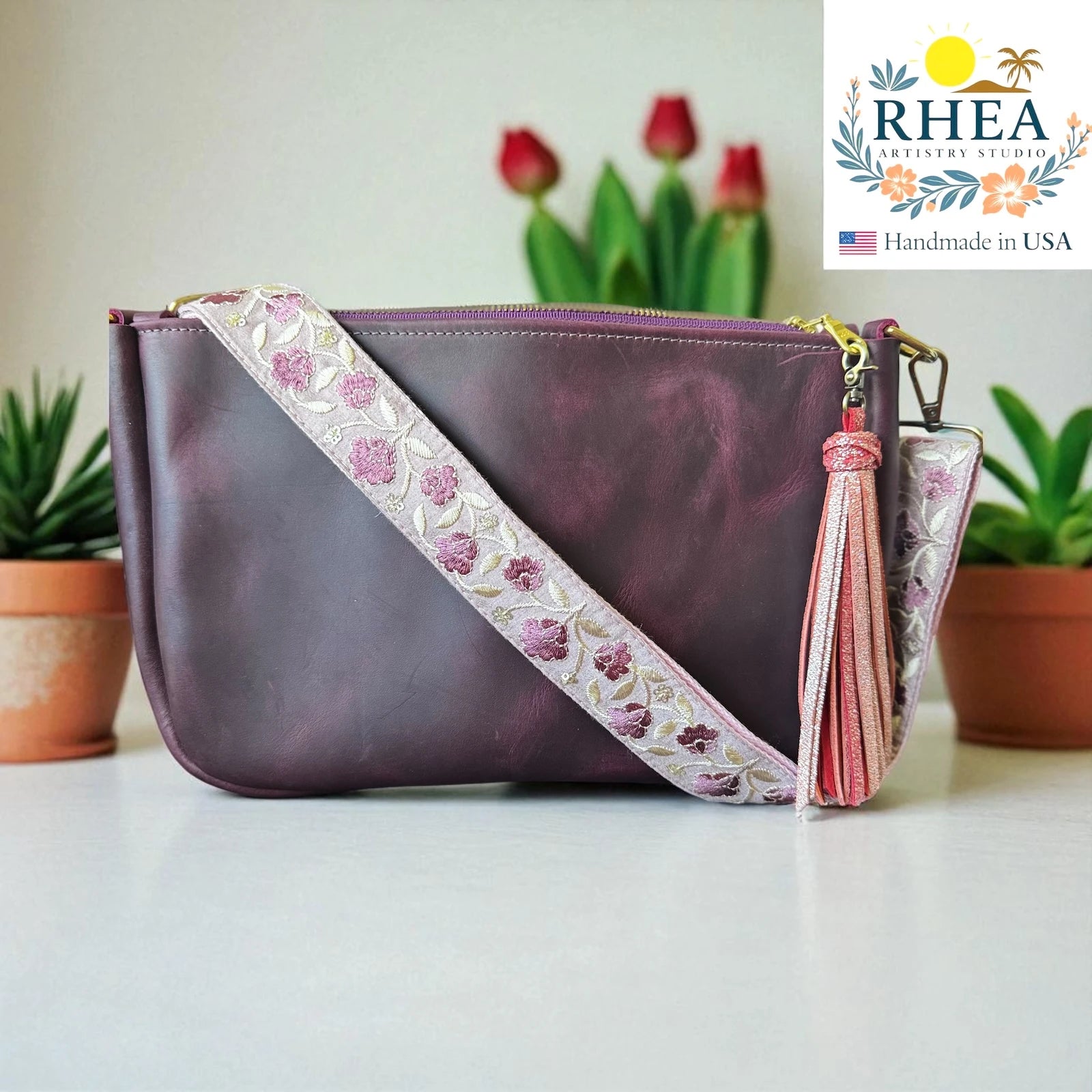 Purple handbag with floral leather purse strap and tassel on a white surface, with 'RHEA' branding in the corner.