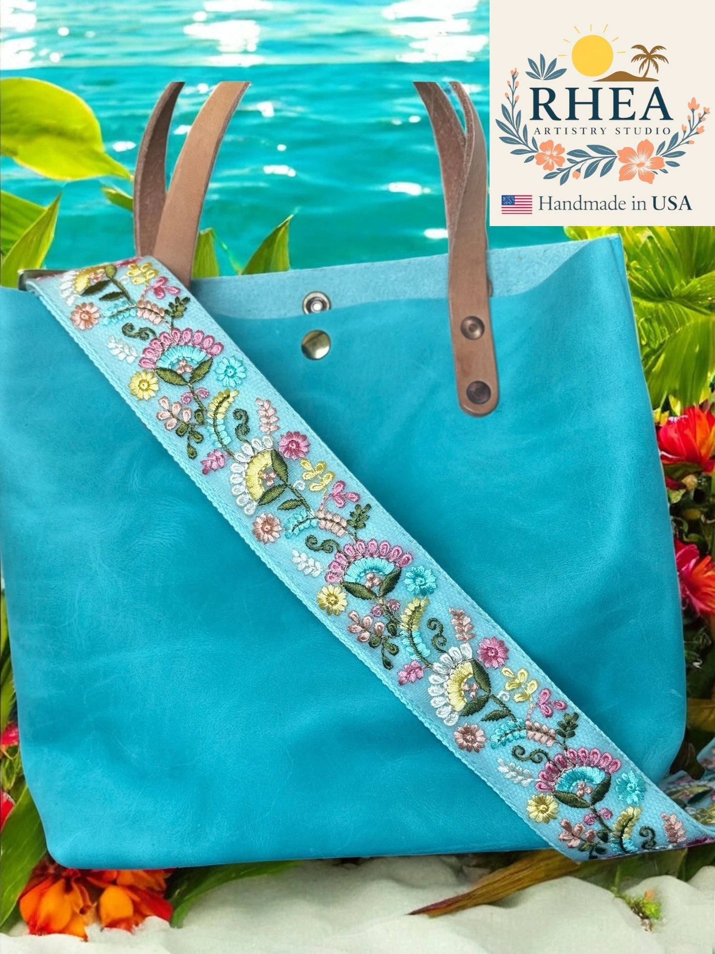 Blue tote bag with floral strap against a natural background, featuring the RHEA brand.