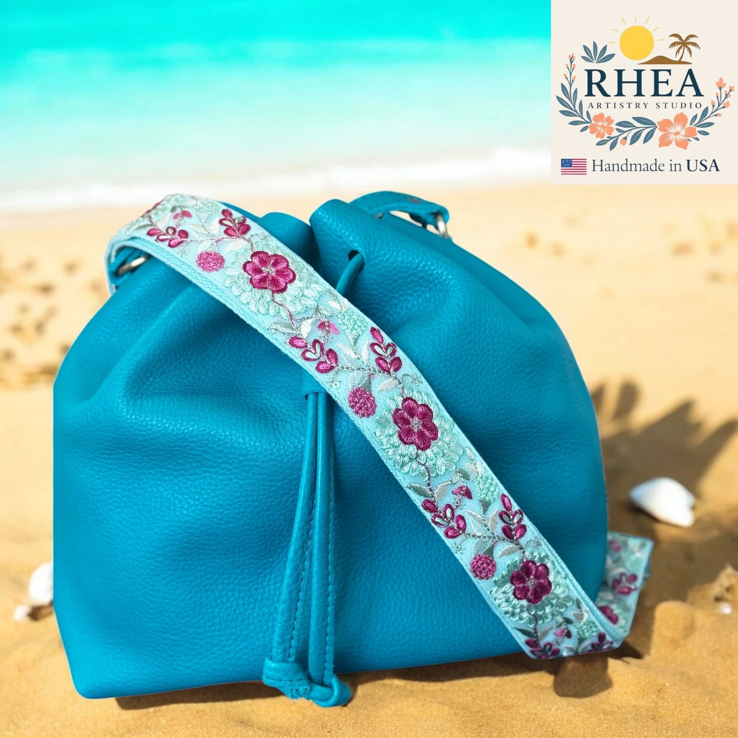 Blue drawstring bag with floral purse strap on a sandy beach, featuring the RHEA brand.