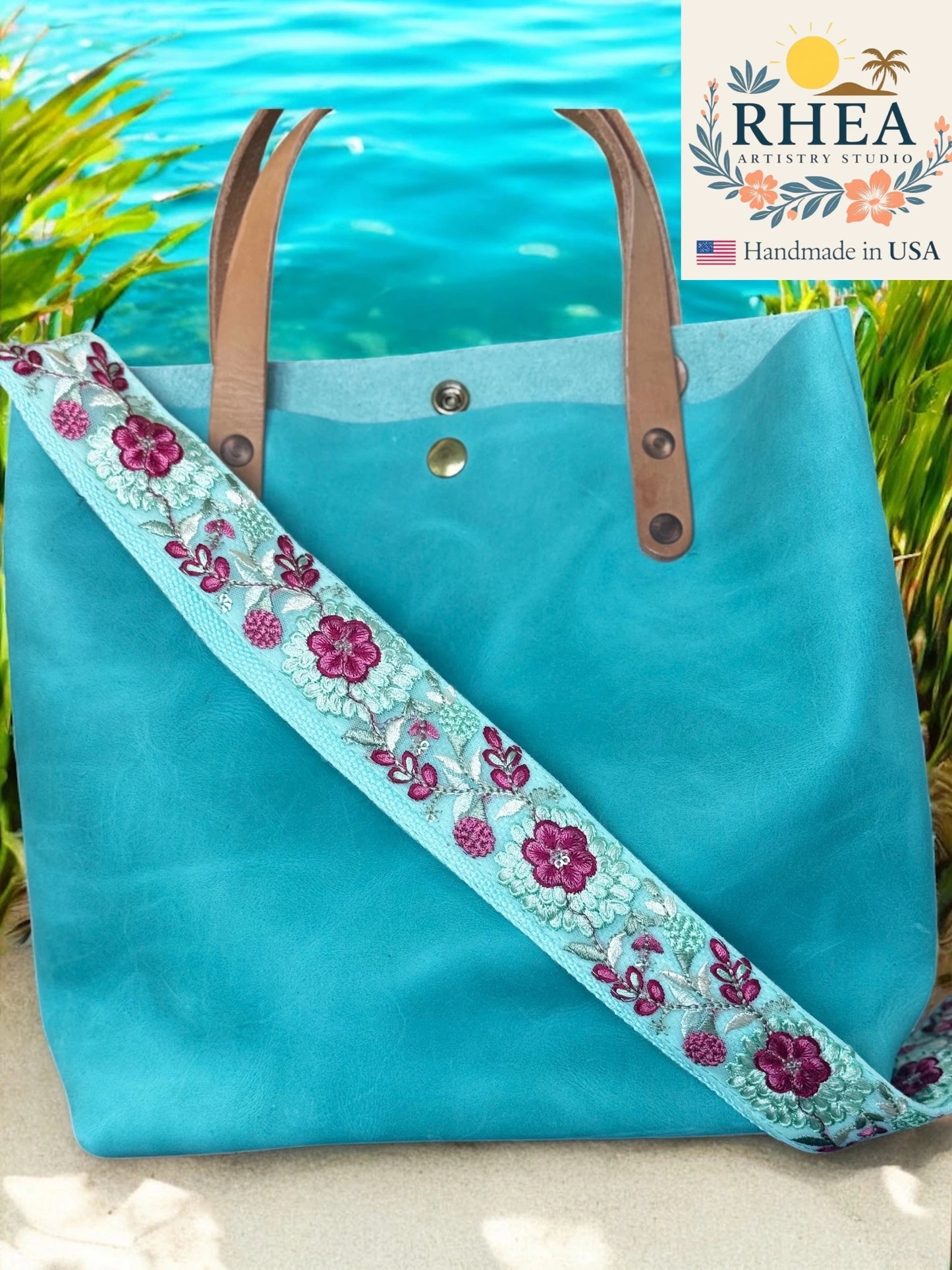 Turquoise tote bag with floral purse strap against a beach background, featuring the RHEA brand.