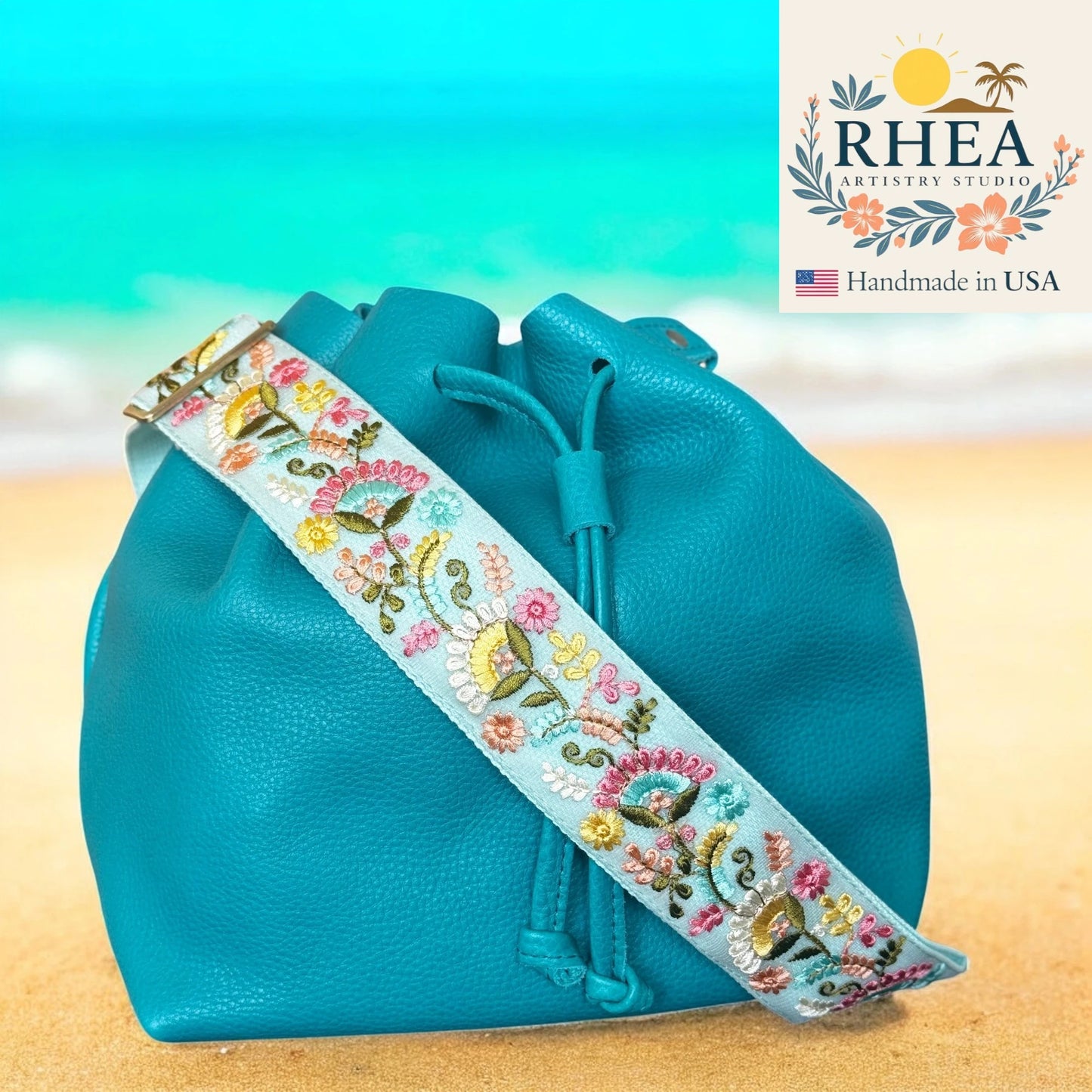 Turquoise drawstring bag with floral embroidery strap on a beach background, featuring the RHEA Artisans Studio logo.