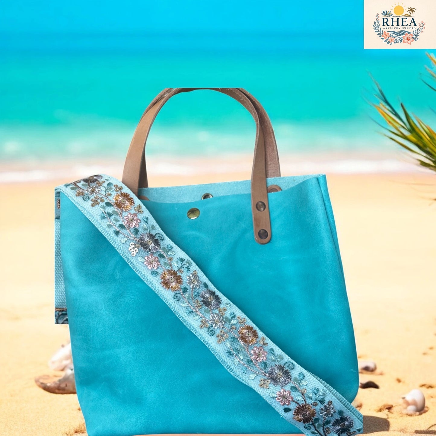 Blue tote bag with floral strap on a beach background, featuring the RHEA brand.