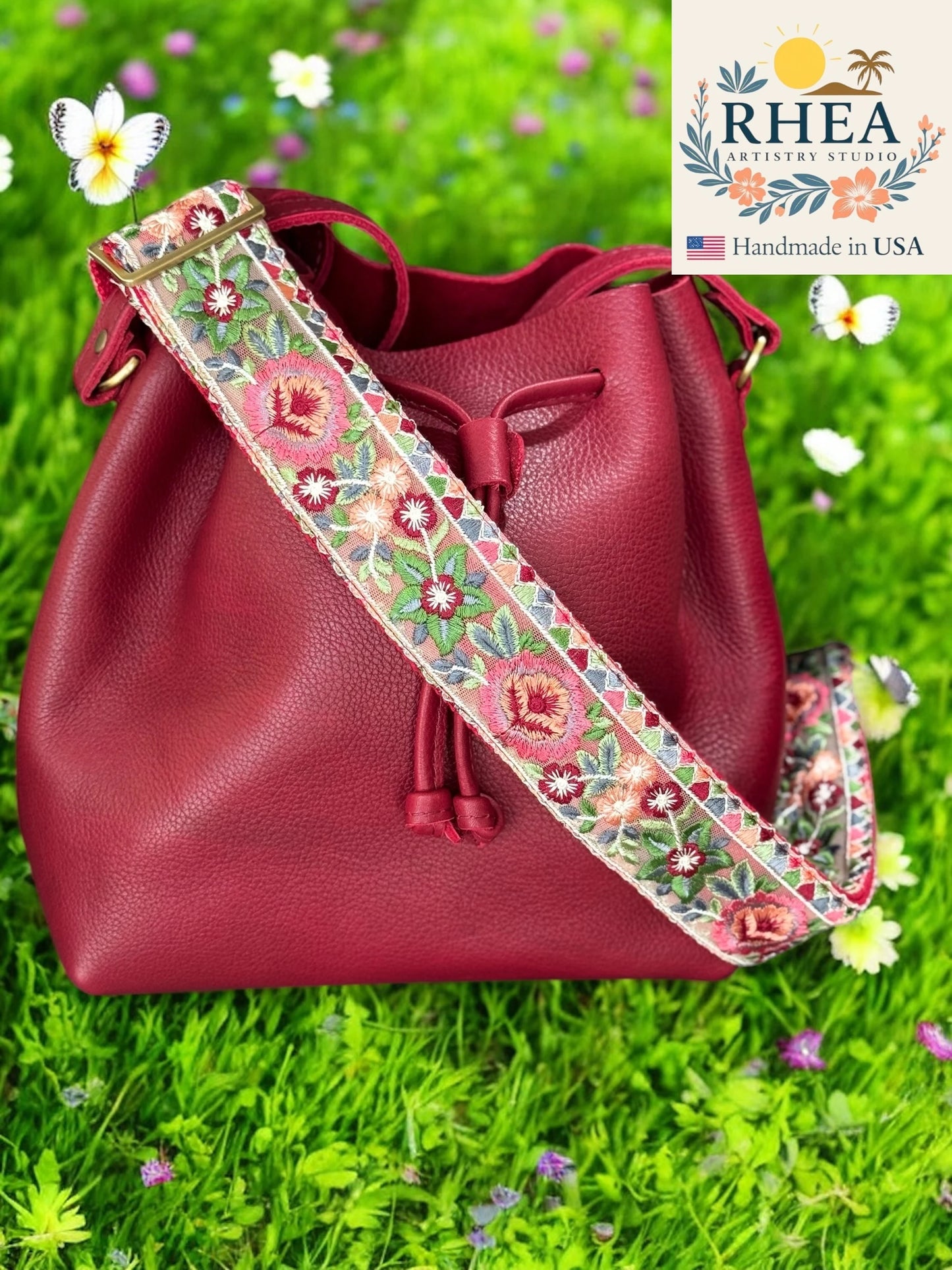 Red handbag with a floral purse strap on grass, featuring the RHEA brand.