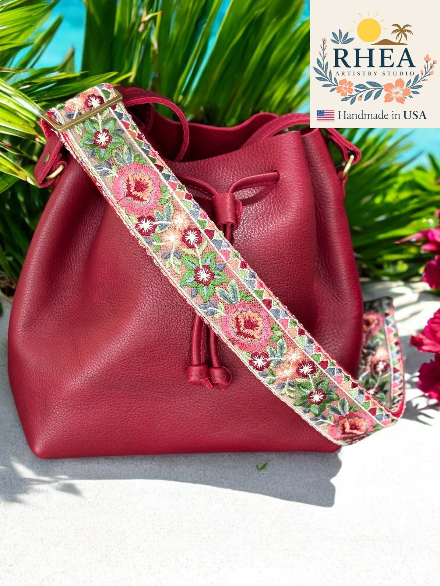 Red leather bucket bag with a floral purse strap against a tropical background, featuring the RHEA brand.