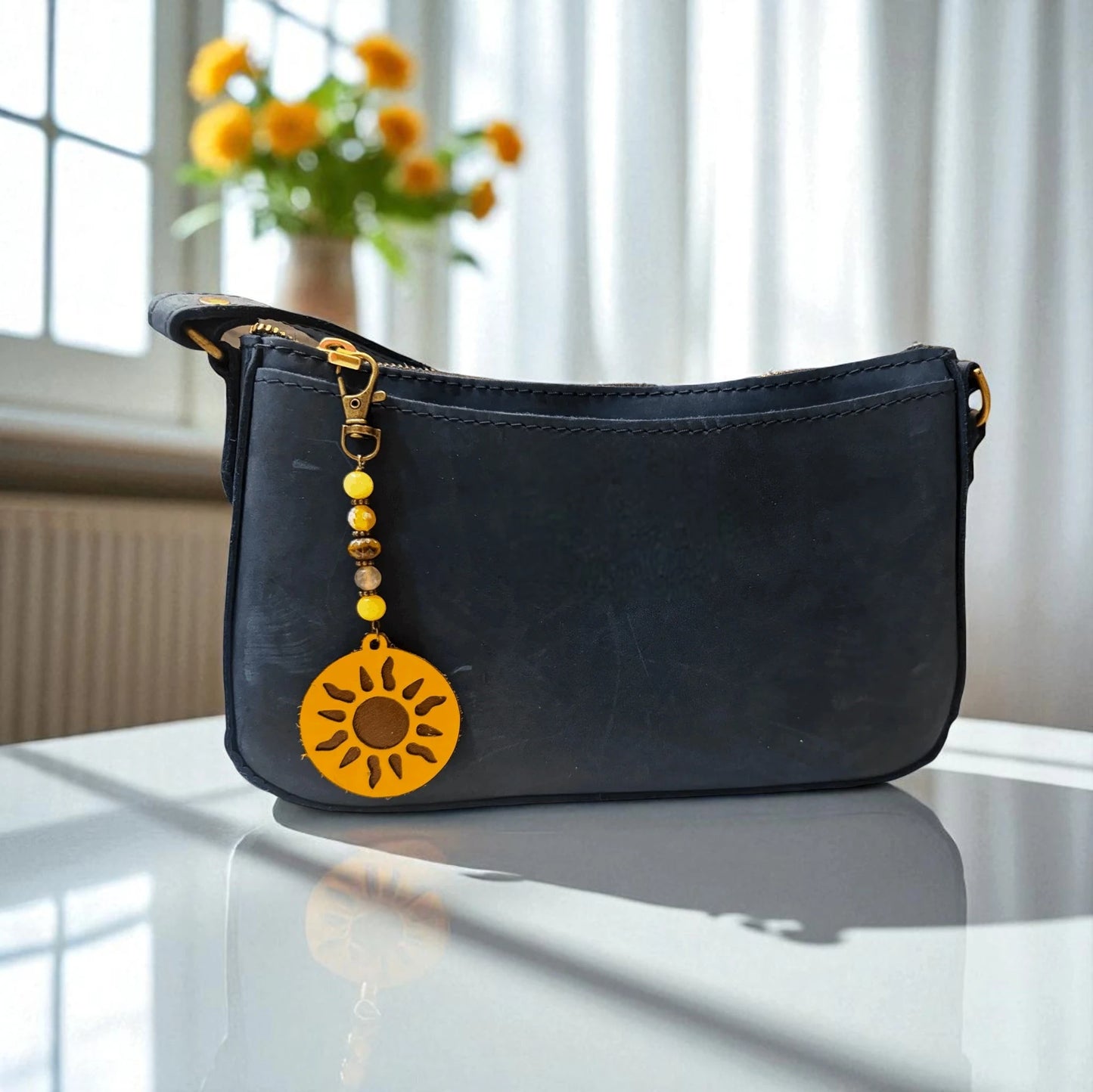 Black handbag with a yellow sun charm on a reflective surface, with a blurred background of flowers and light.