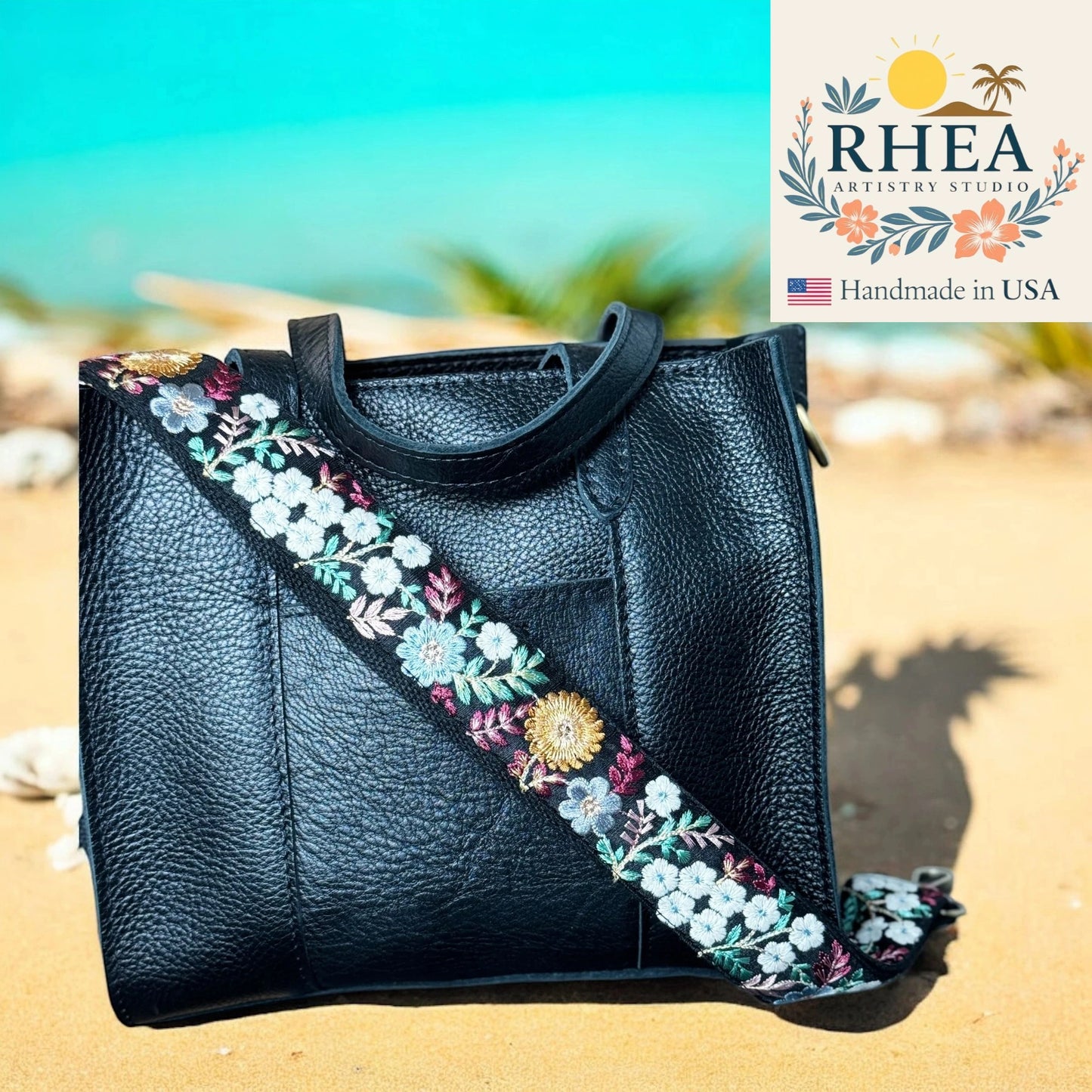 Black handbag with floral strap on a beach, featuring RHEA Artistry Studio logo.