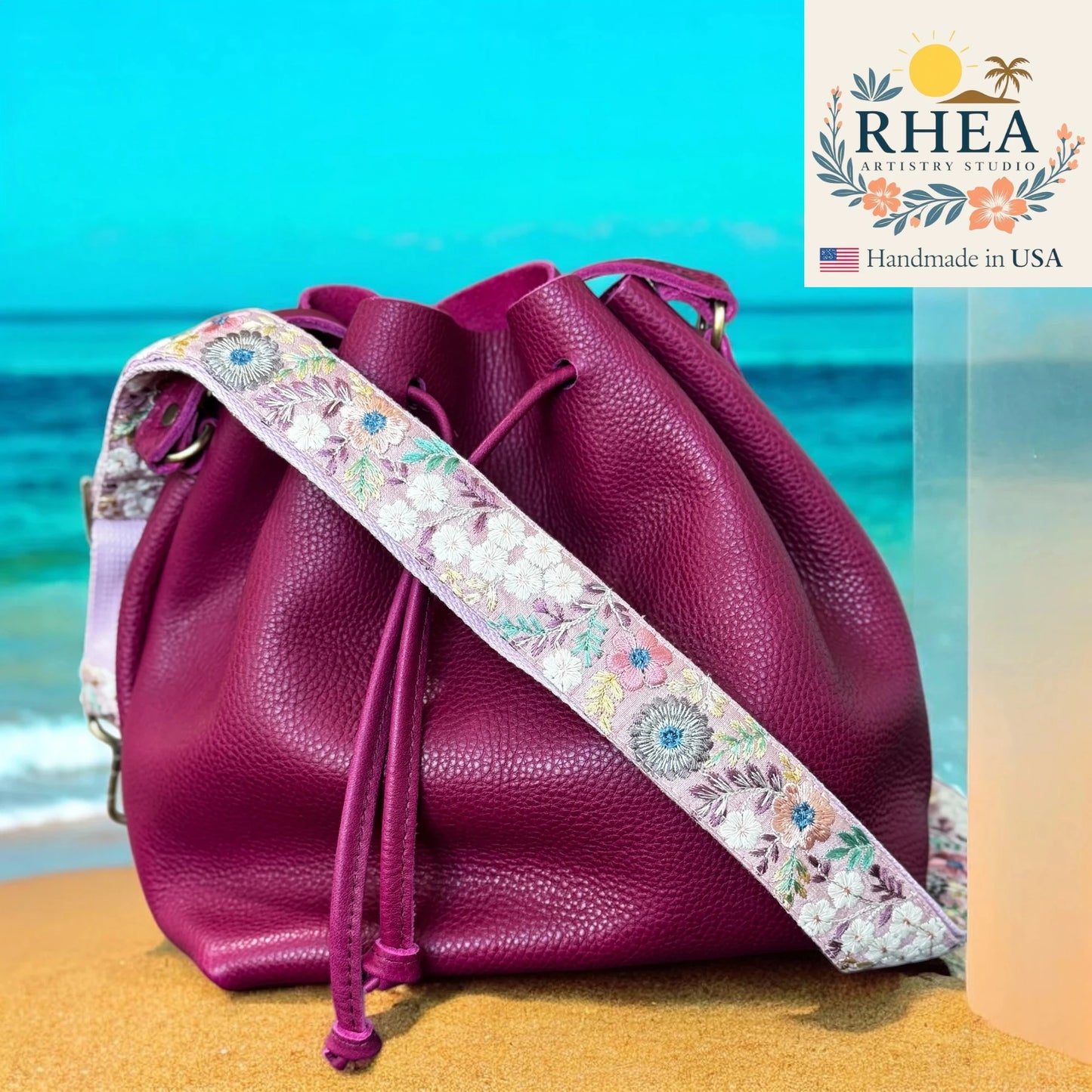 Purple leather bag with a floral strap on a beach background, featuring the Rhea Artistry Studio logo.