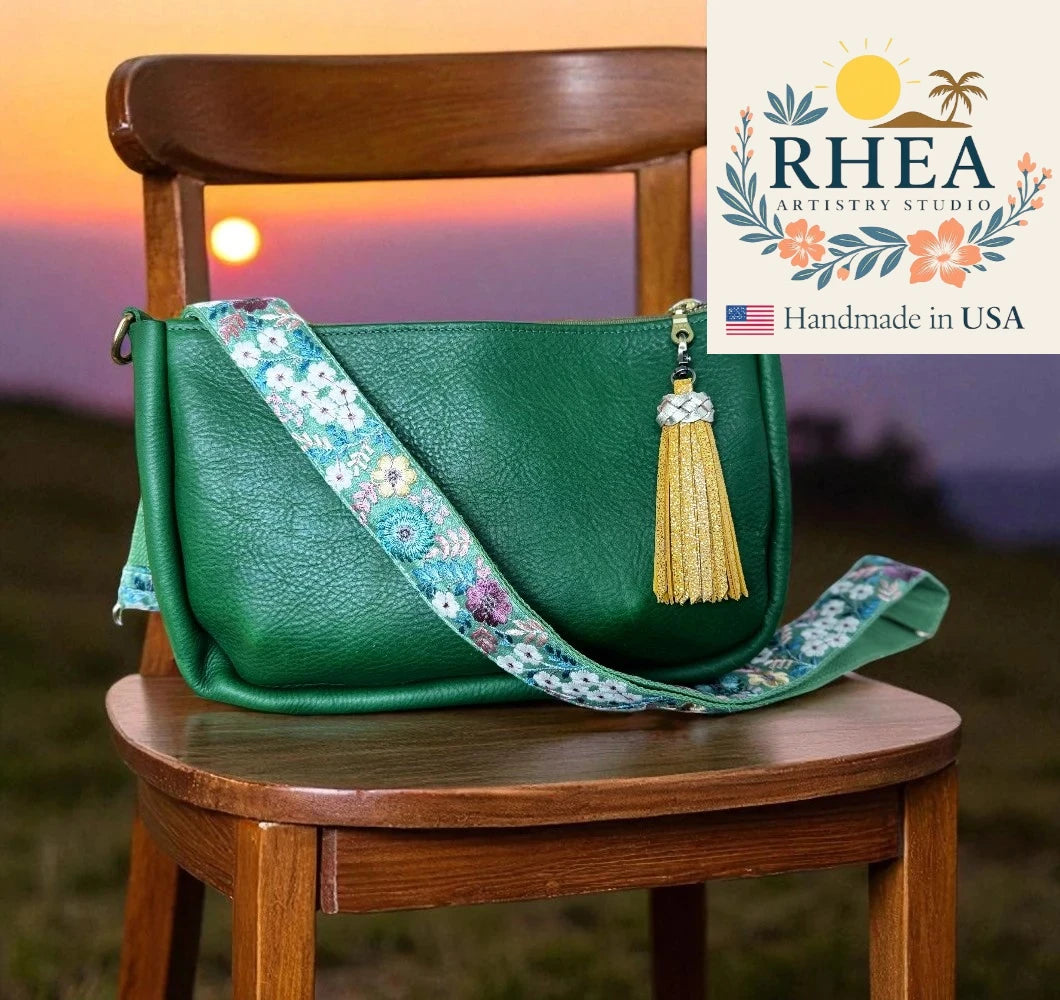 Green handbag with floral strap and tassel on a wooden chair against a sunset background, featuring RHEA Artistry Studio logo.