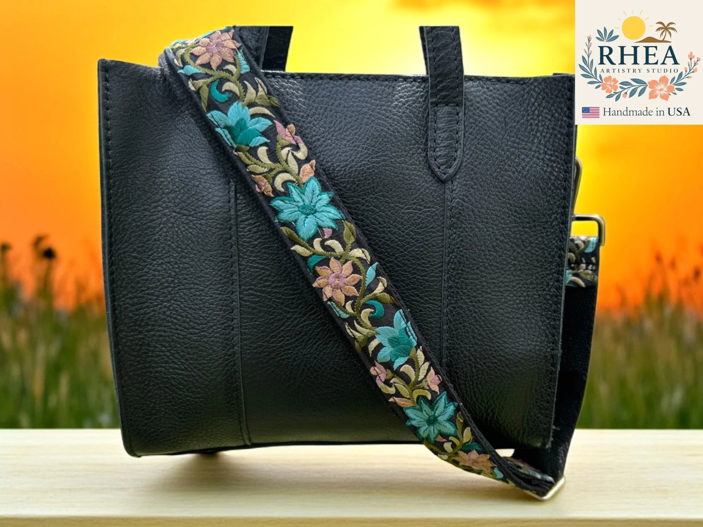 Black leather handbag with a floral purse strap against a blurred natural background, featuring the RHEA brand.