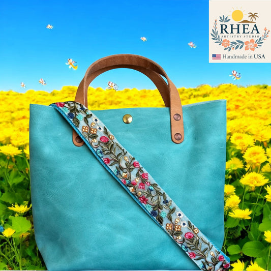 Turquoise tote bag with floral purse strap against a yellow flower field background, featuring the RHEA brand.