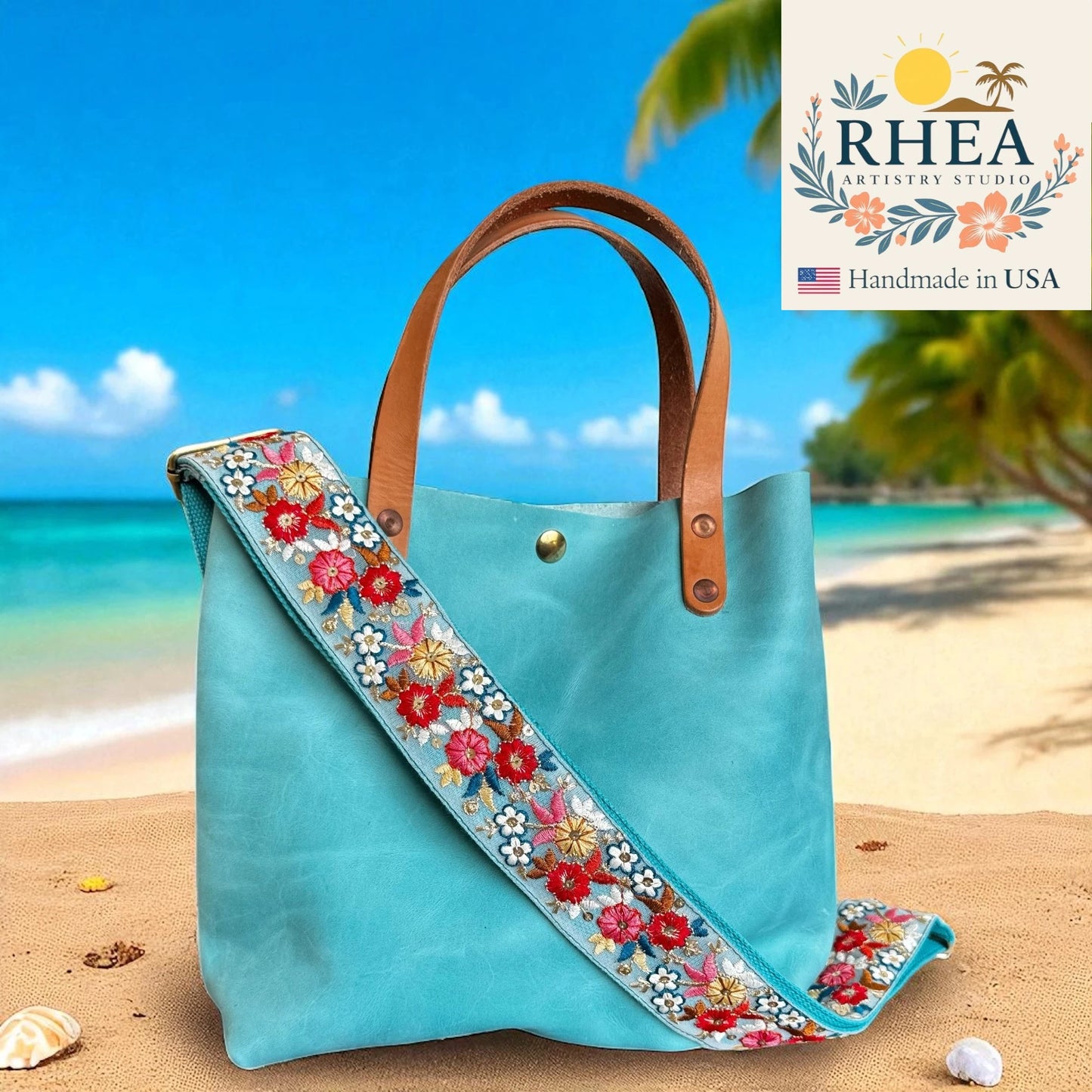 Turquoise tote bag with floral strap on a beach, featuring RHEA Artisans Studio logo.