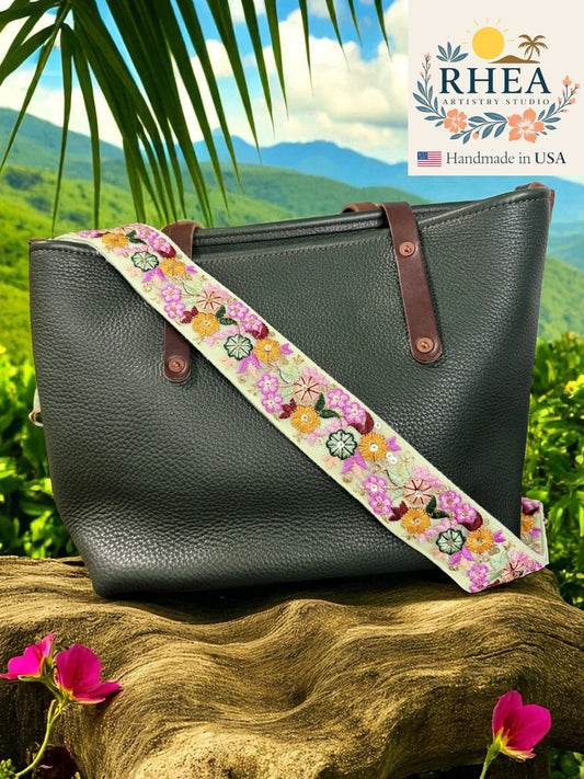 Black handbag with a floral strap on a wooden surface with a scenic background, featuring the RHEA Artistry Studio logo.