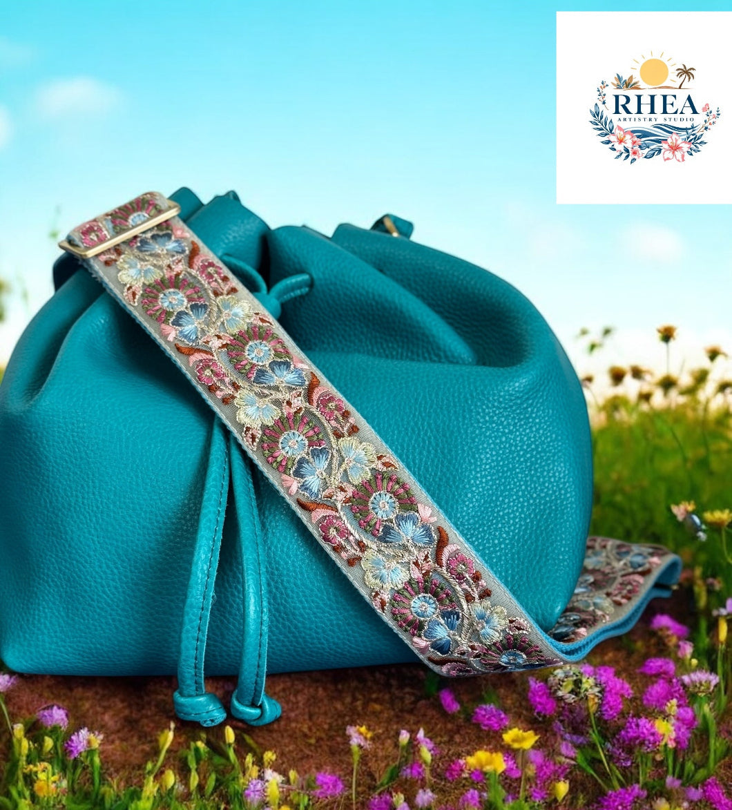 A black purse with a pink and green floral embroidered strap placed on a blue surface with a bright sky and greenery in the background.