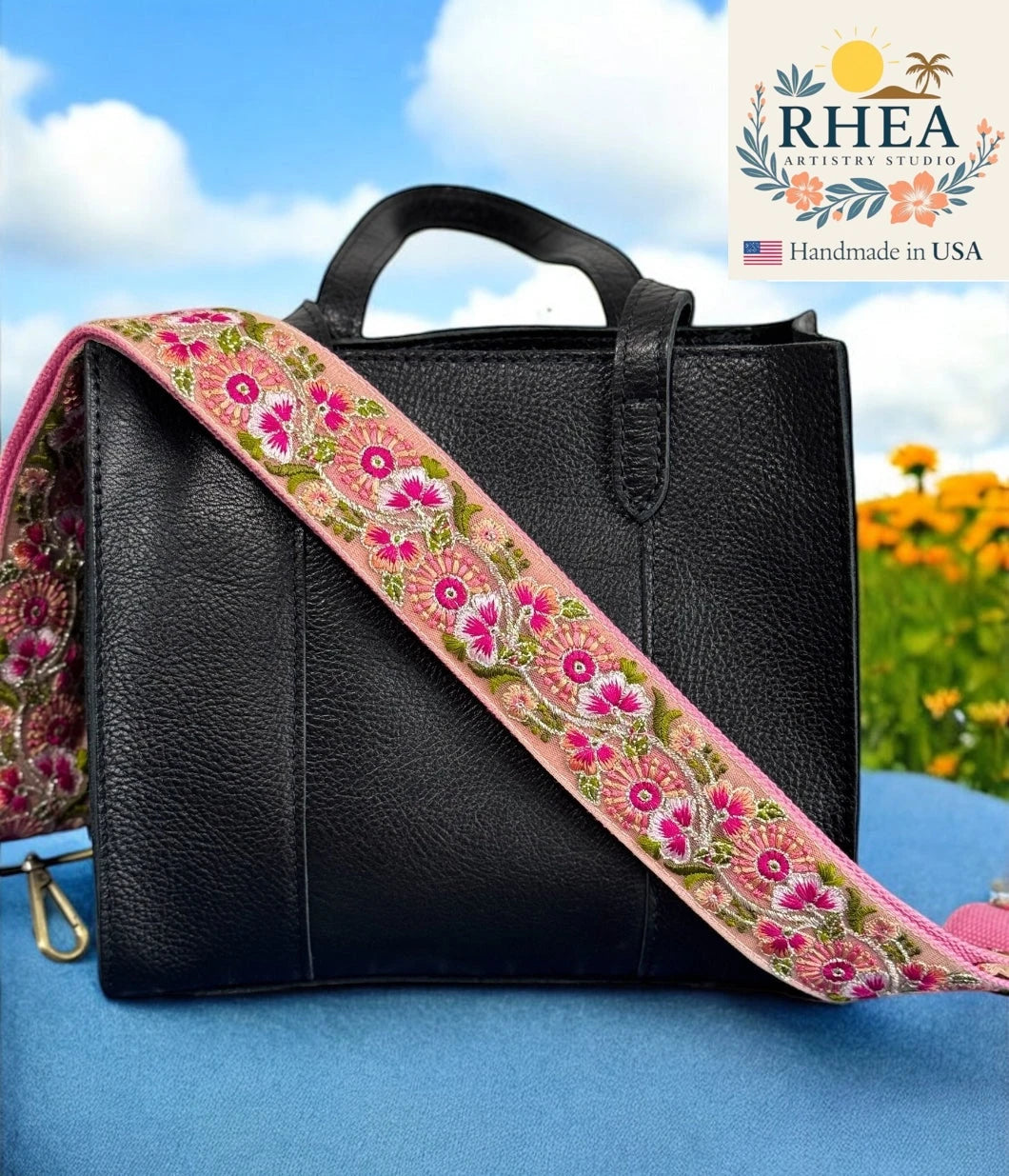 Black leather bag with a colorful floral purse strap against a blue sky and flower background, featuring the RHEA brand.
