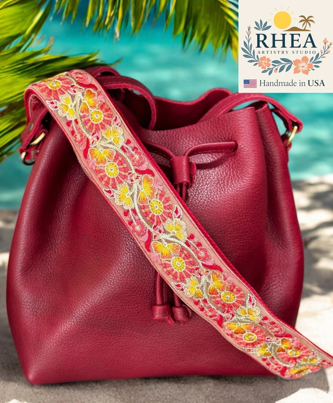 Red leather bag with a floral purse strap on a beach background, featuring the RHEA brand.
