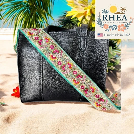 Black handbag with a colorful floral purse strap on a beach background, featuring the RHEA brand.