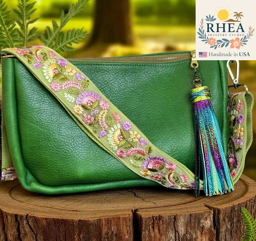 Green handbag with floral strap and tassel on a wooden surface, RHEA brand visible.