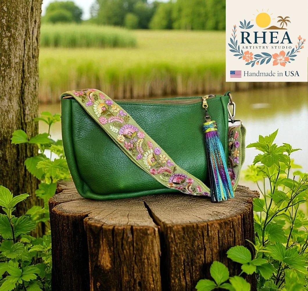Green handbag with floral strap on a wooden stump in a natural setting, featuring RHEA Artistry Studio branding.