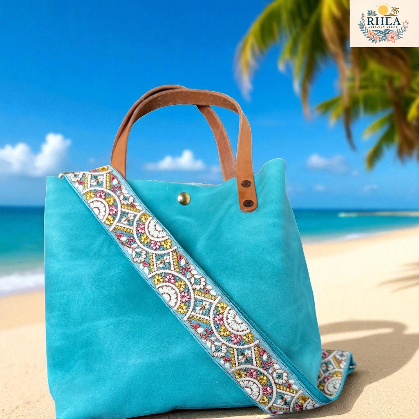 Turquoise tote bag with a colorful strap on a beach, featuring the RHEA brand.