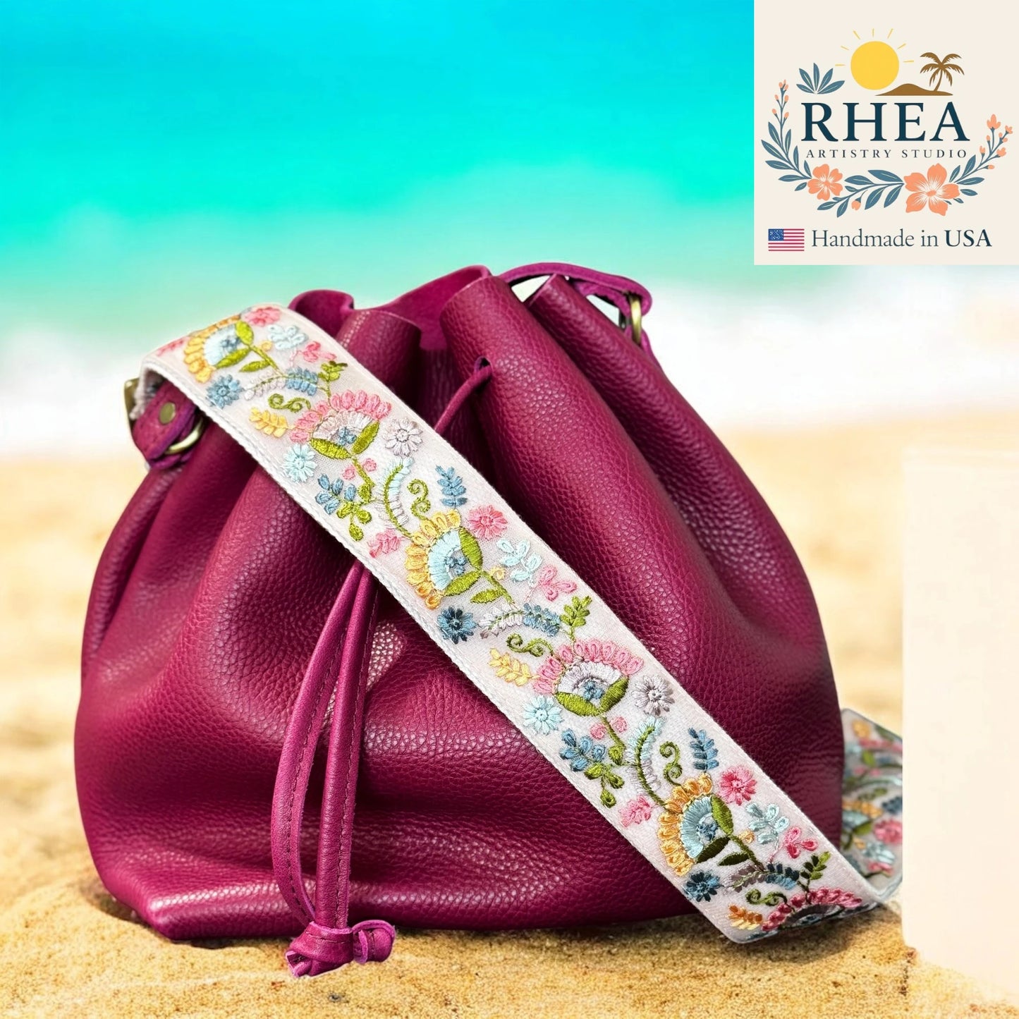 Pink leather bag with an embroidered strap on a sandy background, featuring the Rhea Artistry Studio logo.