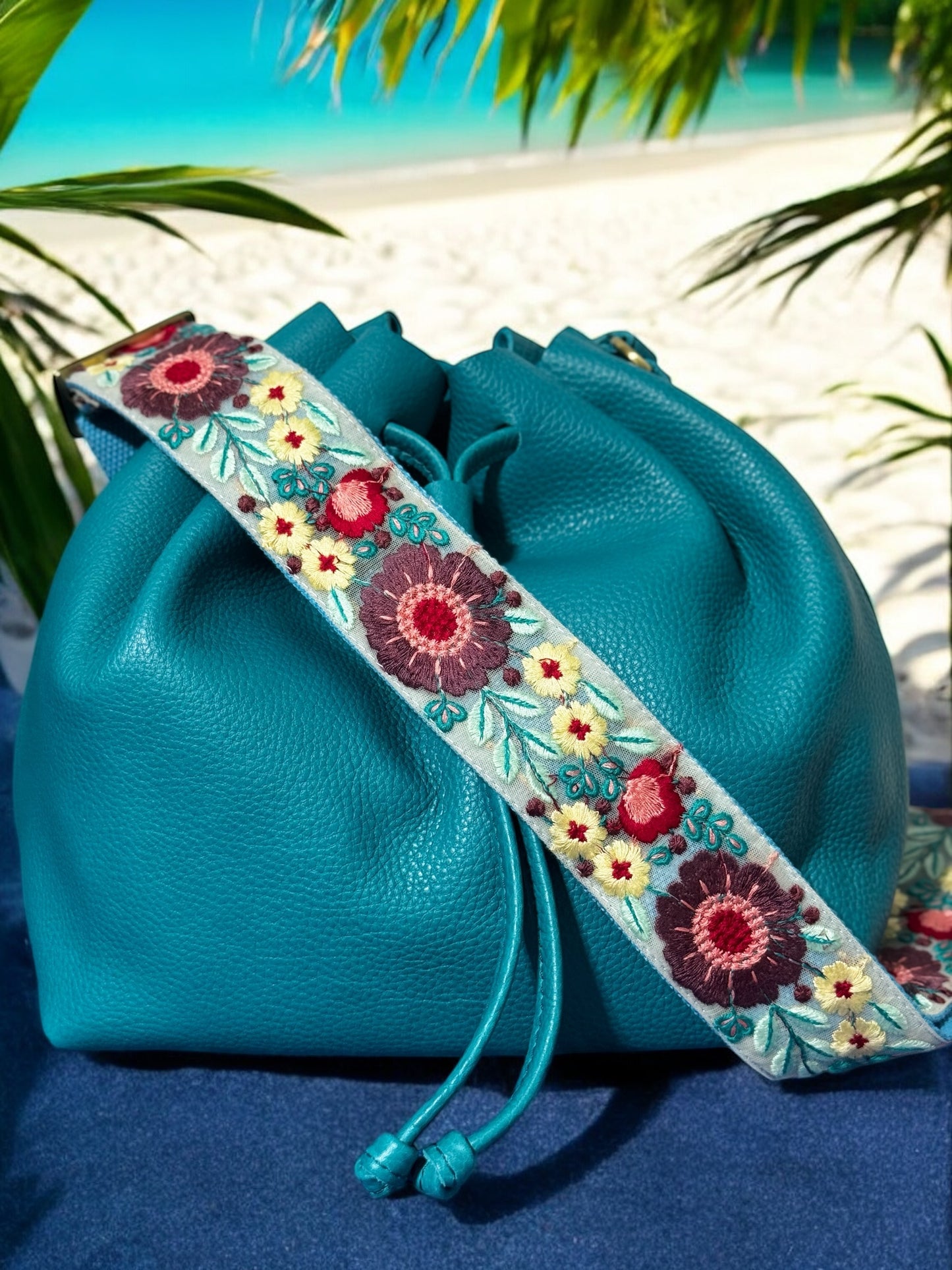 Vibrant turquoise purse strap featuring large blue and yellow floral embroidery, styled on a deep navy drawstring leather bag.