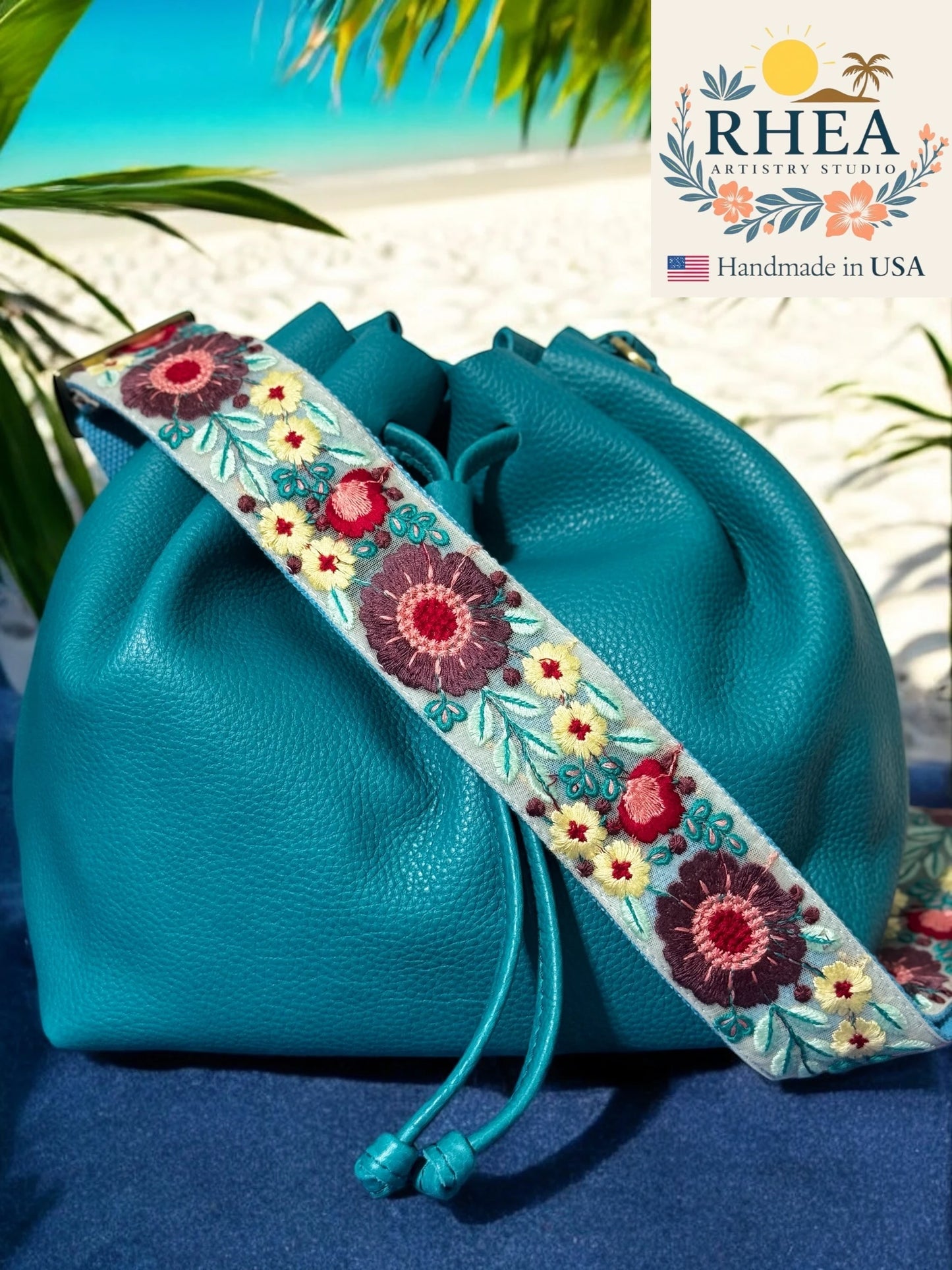 Turquoise handbag with a floral purse strap on a beach background, featuring the RHEA brand.