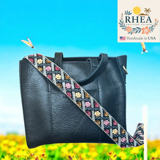 Black handbag with a colorful strap against a blue sky and yellow flower background, featuring the Rhea Artistry Studio logo.