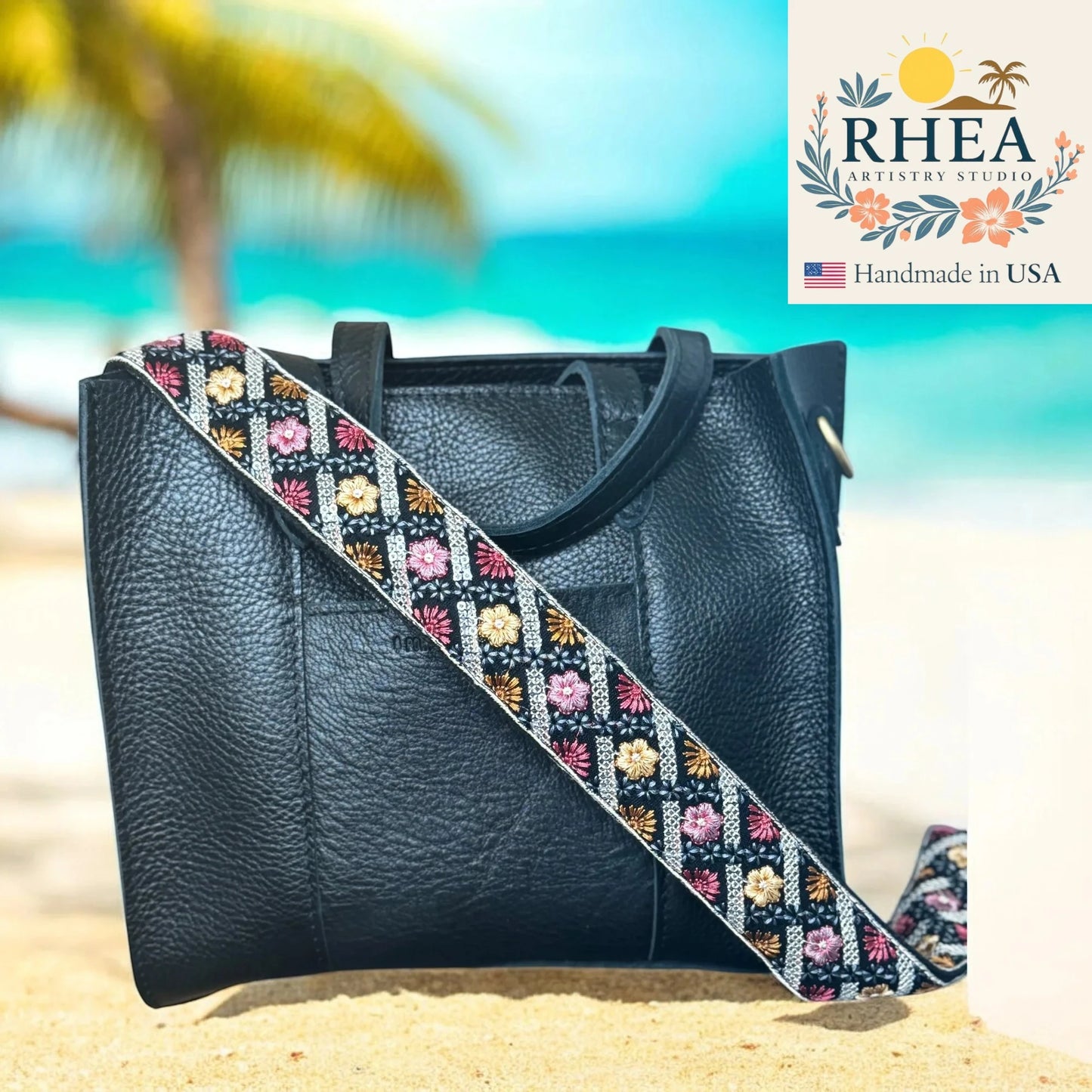 Black leather handbag with a colorful strap on a beach, featuring the RHEA Artisan Studio logo.