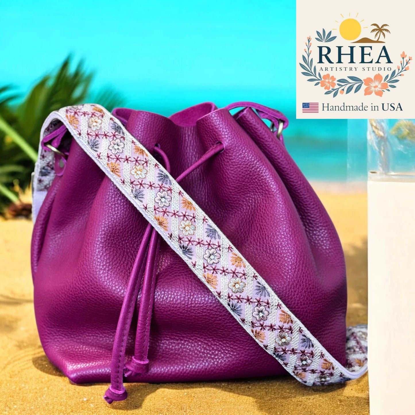 Purple leather bag with a floral strap on a beach, featuring the Rhea Artistry Studio logo.