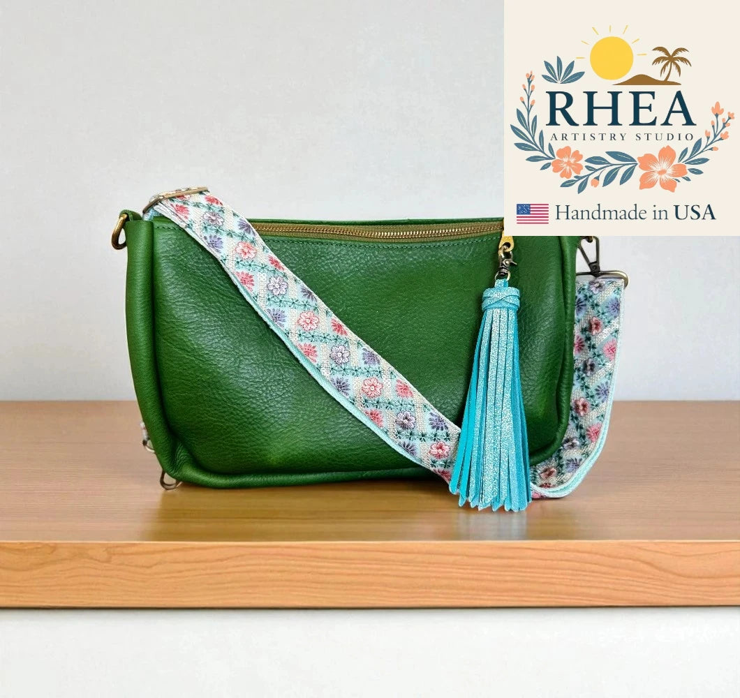 Green handbag with floral strap and tassel on a wooden surface, RHEA Artistry Studio logo visible.