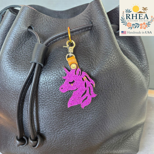 Black leather bag with a purple unicorn keychain charm attached, featuring the RHEA brand logo.