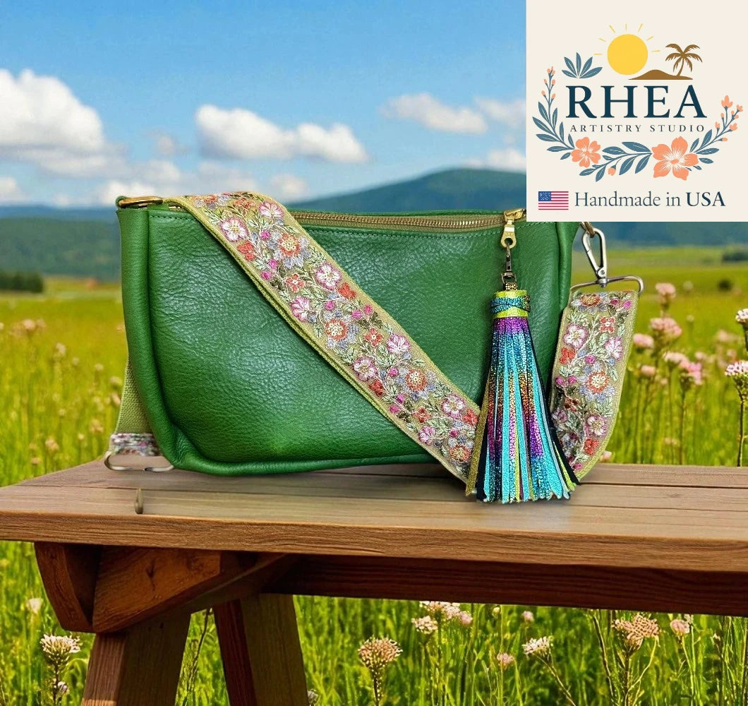 Green handbag with floral strap and tassel on a wooden table in a field with 'RHEA Artistry Studio' branding.