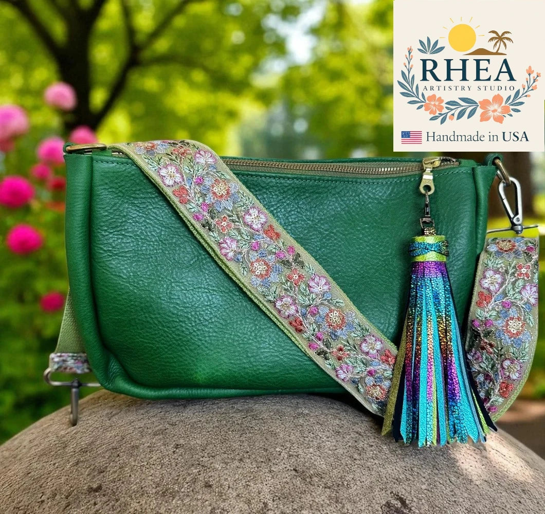Green handbag with floral strap and tassel on a rock with blurred flowers and trees in the background, RHEA Artisan Studio logo visible.