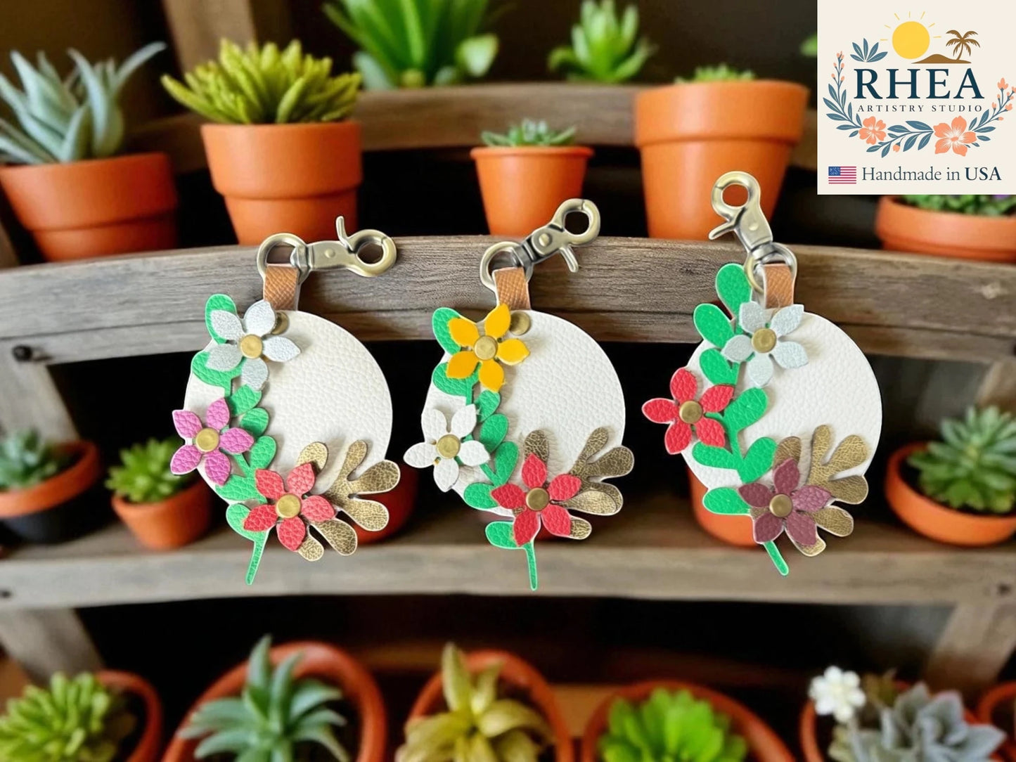 Floral leather bag charms on a wooden shelf with potted plants in the background, featuring the RHEA brand.