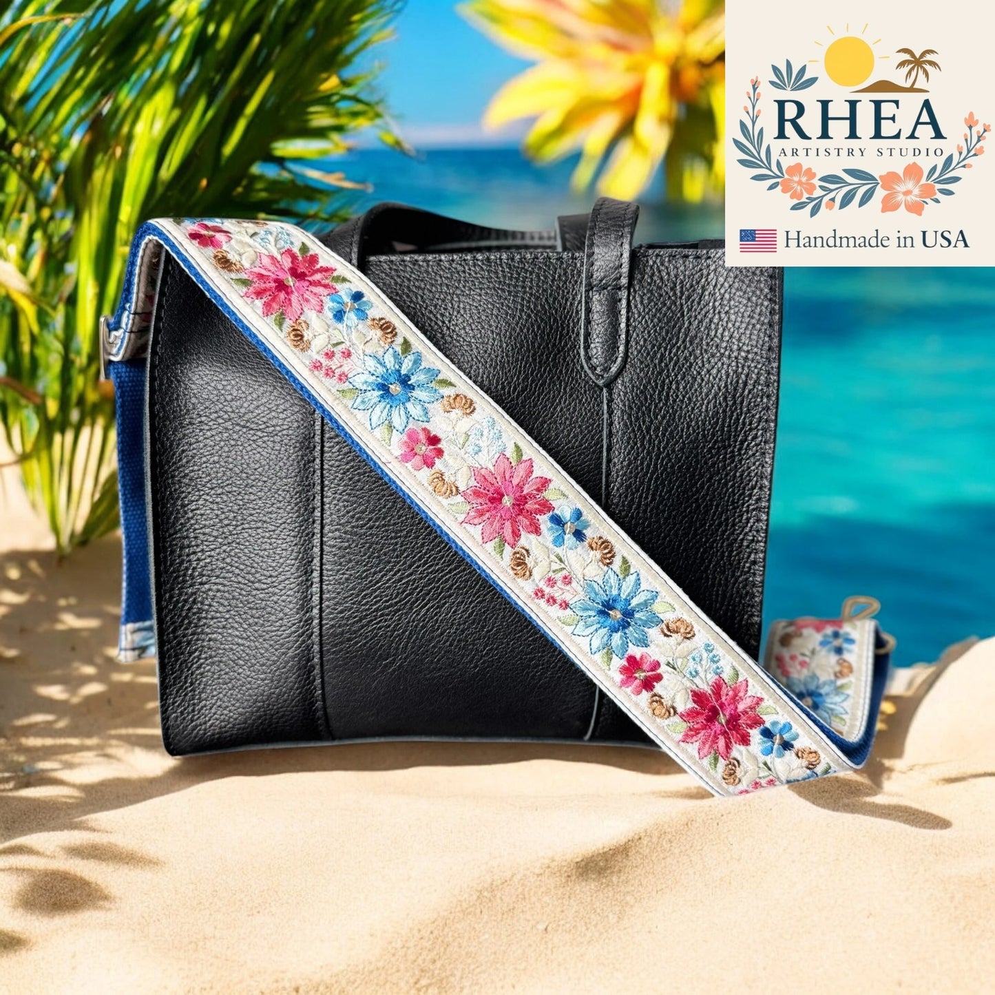 Black handbag with a floral purse strap on a sandy beach with palm trees and blue water.